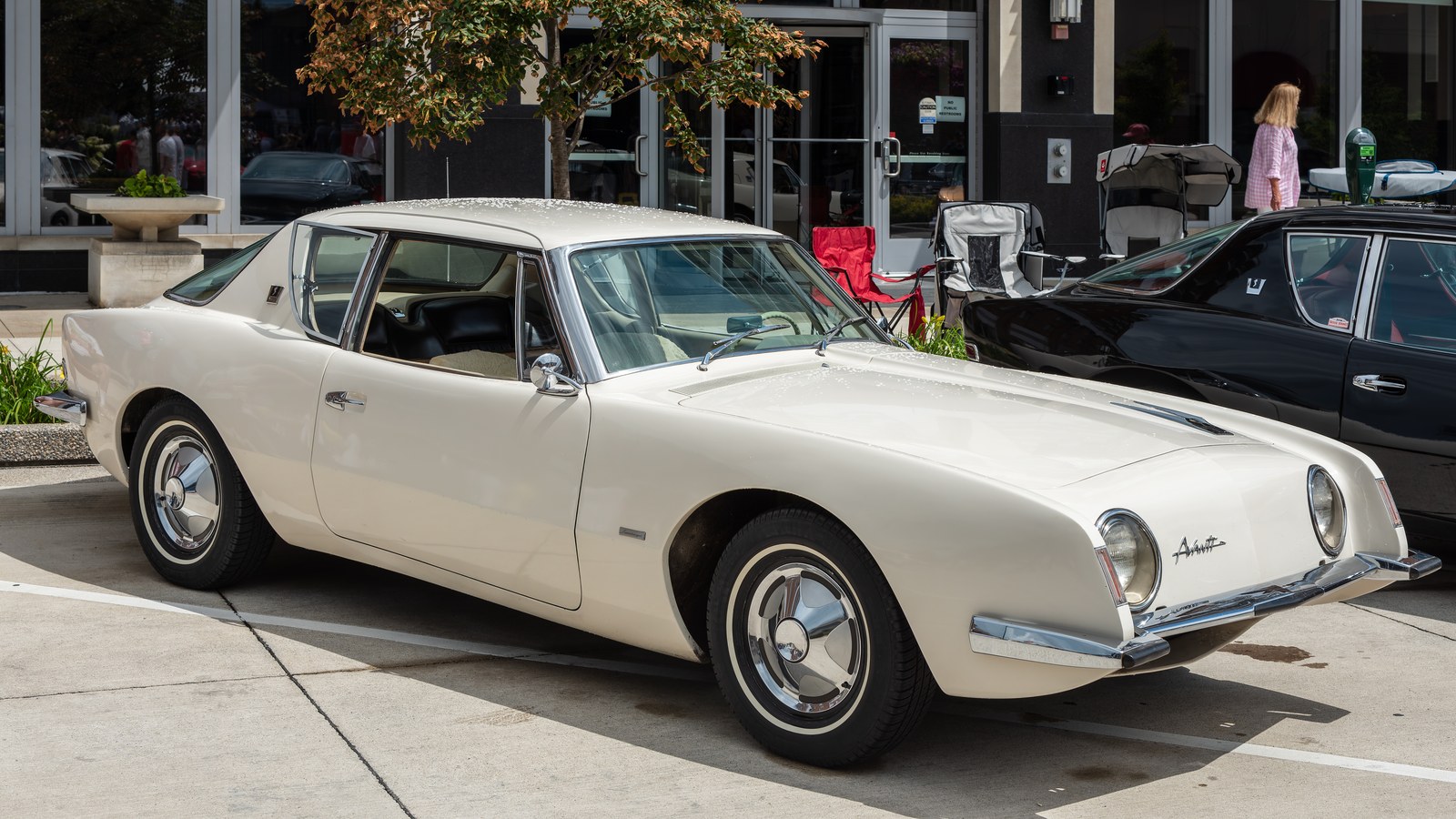 Un Studebaker Avanti 1963 color blanco marfil, visto de lado desde la derecha, estacionado sobre concreto frente a un edificio con ventanales, con un auto negro y sillas plegables detrás.