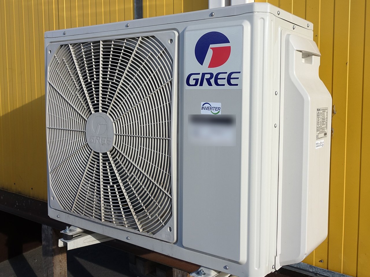 Unidad exterior blanca de una bomba de calor tipo inverter, con un ventilador grande, montada sobre soportes de metal junto a una pared amarilla.
