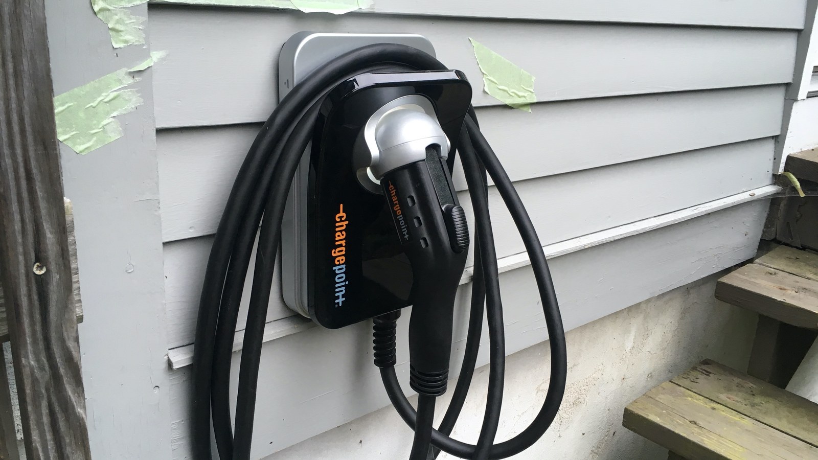 Un cargador de nivel 2 para auto eléctrico, de color negro, instalado en la pared exterior de una casa de madera gris, con el cable enrollado alrededor del soporte.