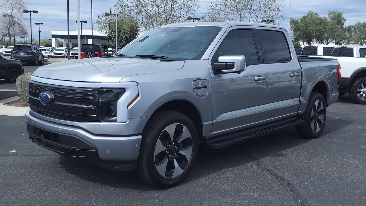 Ford F-150 Lightning Platinum 2023 color plata, cabina doble, vista delantera de tres cuartos, con la barra de luz que cruza todo el frente, la parrilla cerrada y el óvalo azul de Ford, estacionada en el lote de un concesionario.