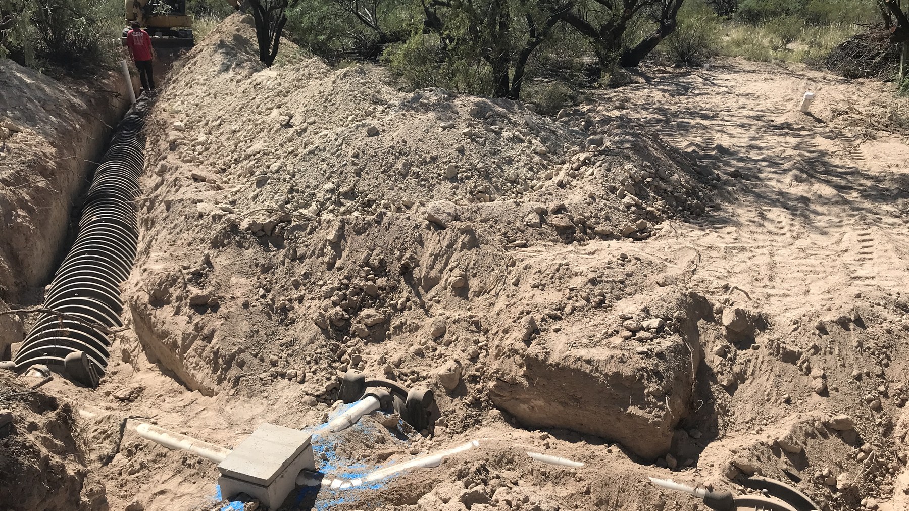 Obra de instalación de un sistema séptico residencial: zanja con cámara negra corrugada de campo de absorción, caja de distribución de concreto con tubería PVC blanca y excavadora al fondo en terreno arenoso.