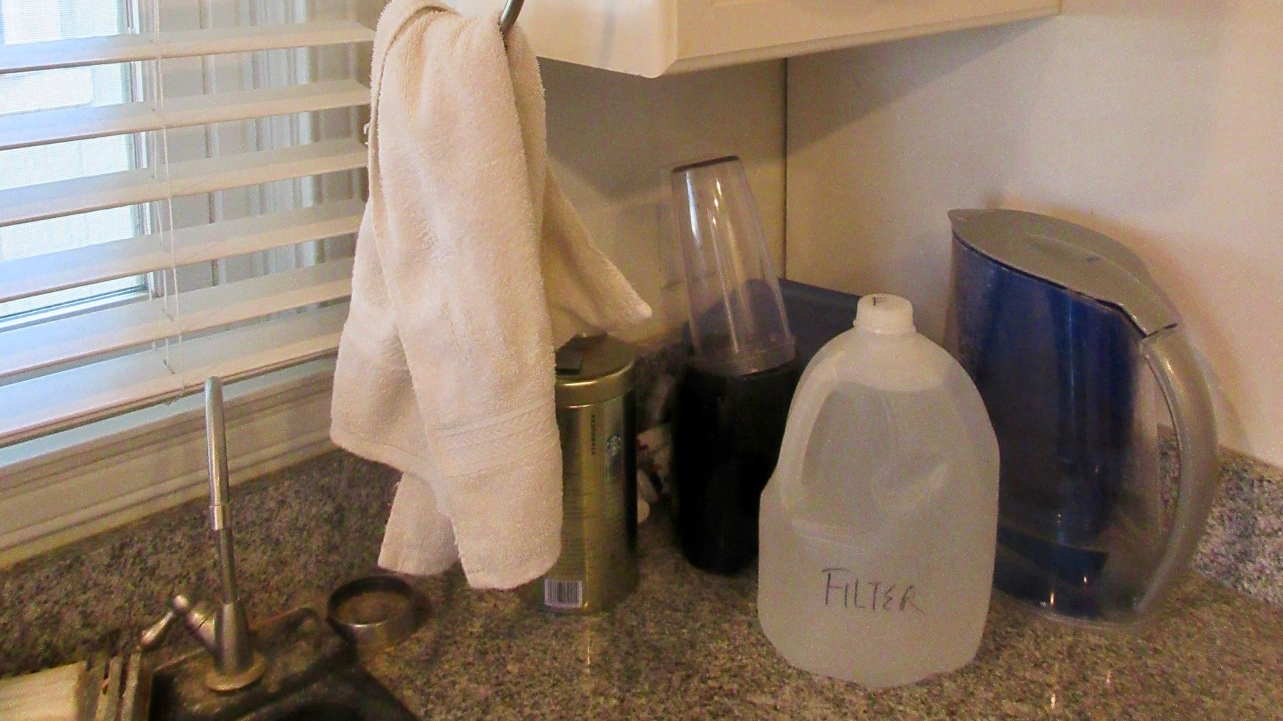 Rincón de una cocina residencial con un grifo auxiliar delgado junto al fregadero, una jarra filtrante azul y un galón de plástico marcado FILTER sobre la cubierta de granito.