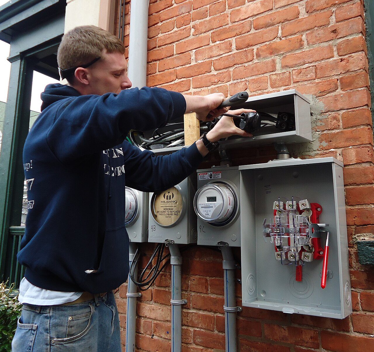 Un electricista trabaja con un destornillador en los medidores y la caja de breakers en la pared de una casa.