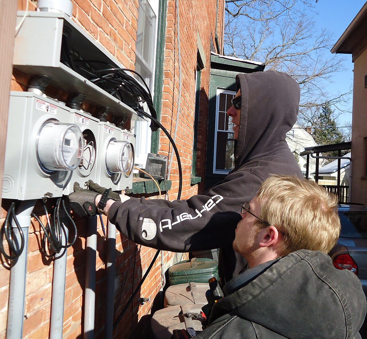 Dos electricistas instalan medidores de luz en la pared de ladrillo de una casa de varias familias.